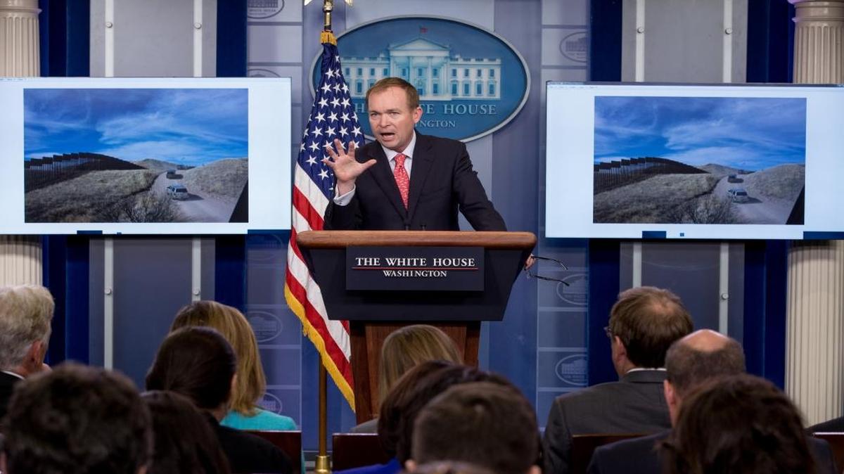 An image depicting a section of border wall is displayed behind Budget Director Mick Mulvaney as he speaks to the media during the daily press briefing at the White House.